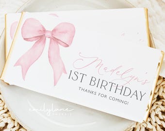 May include: A white chocolate bar wrapper with a pink watercolor bow and the text "Madelyn's 1st Birthday Thanks for coming!"  The wrapper is decorated with a pink watercolor bow and the text "Madelyn's 1st Birthday Thanks for coming!"  The wrapper is designed by Emily Jane Paperie.