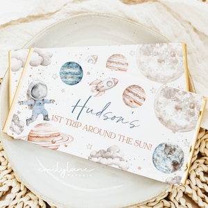 Puede incluir: Un envoltorio de chocolate blanco con temática espacial. Presenta un astronauta de dibujos animados, planetas, estrellas y nubes. El texto en el envoltorio dice "Hudson's 1st Trip Around The Sun!" y "emilylane paperie".