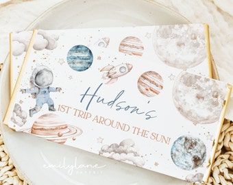 May include: A white chocolate bar wrapper with a space theme. It features a cartoon astronaut, planets, stars, and clouds. The text on the wrapper reads "Hudson's 1st Trip Around The Sun!" and "emilylane paperie".