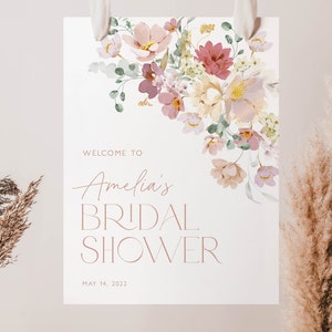 May include: A white bridal shower sign with a floral design in shades of pink, purple, and yellow. The text reads "Welcome to Amelia's Bridal Shower May 14, 2022".