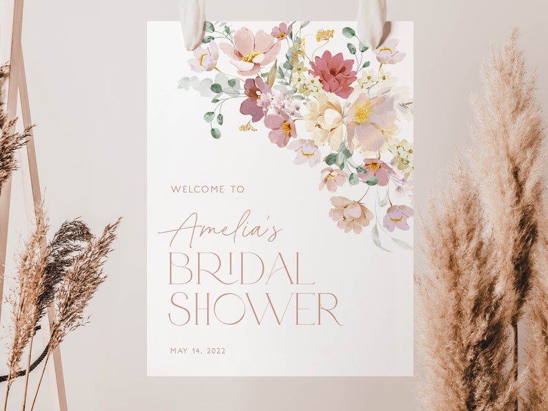 May include: A white bridal shower sign with a floral design in shades of pink, purple, and yellow. The text reads "Welcome to Amelia's Bridal Shower May 14, 2022".