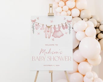 May include: A welcome sign for a baby shower, decorated in pink and white. The sign features a clothesline with baby clothes hanging on it, including a pink dress, a pink onesie, and a pair of pink shoes. The text on the sign reads "Welcome to Madison's Baby Shower, December 4, 2023".