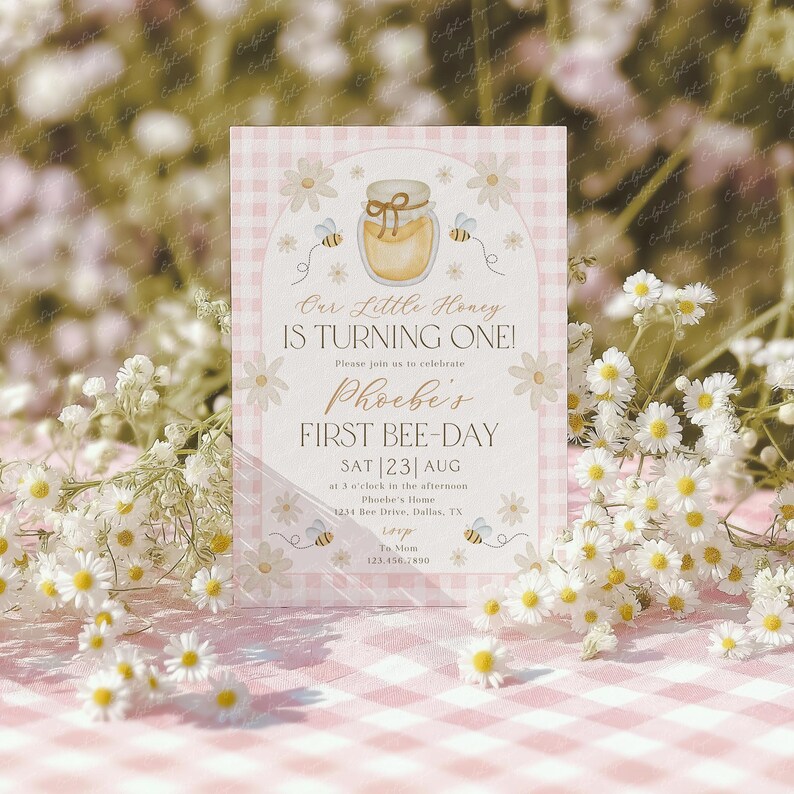 May include: A pink and white invitation card with a bee theme. The card reads "Our Little Honey Is Turning One!" and "Phoebe's First Bee-Day" with the date and time. The card is decorated with daisies, bees, and a jar of honey.