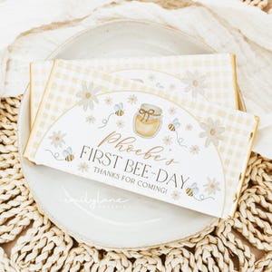 Puede incluir: Dos recuerdos de fiesta rectangulares con un patrón de cuadros amarillos y detalles florales, con el texto "Phoebe's First Bee-Day". Los recuerdos incluyen ilustraciones de abejas y un tarro de miel, sobre un plato blanco.