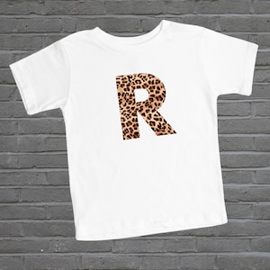May include: White t-shirt with a large letter "R" printed on the front. The letter is made of a leopard print pattern.