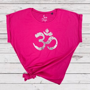 以下が含まれることがあります： 前面に銀色のオームのシンボルがプリントされたピンクのTシャツ。ゆったりとしたフィット感で、肩が少し落ちています。ブランド名は「インカデザイン」とタグに書かれています。