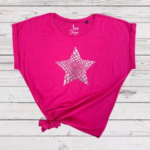Pu&ograve; includere: Una maglietta rosa con una stampa a stella argentata. La stella ha un motivo a pelle di serpente. La maglietta ha un collo rotondo e maniche corte.