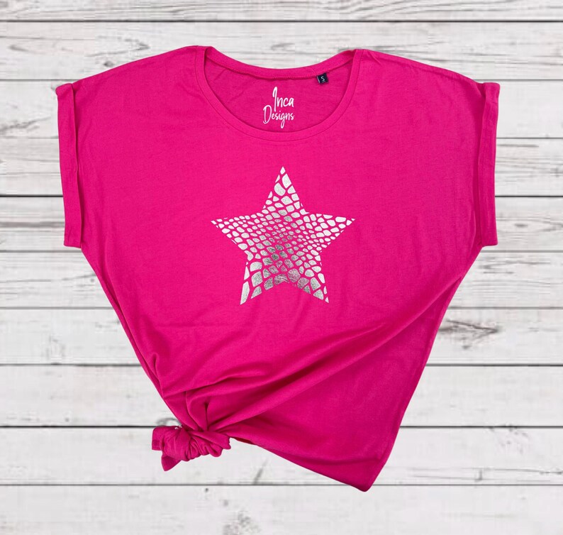 Pu&ograve; includere: Una maglietta rosa con una stampa a stella argentata. La stella ha un motivo a pelle di serpente. La maglietta ha un collo rotondo e maniche corte.