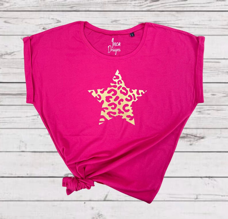 Pu&ograve; includere: Una maglietta rosa brillante con una stella dorata con un motivo a stampa leopardata. La maglietta ha una vestibilit&agrave; rilassata e un orlo annodato. Il testo "Inca Designs" &egrave; stampato sul davanti della maglietta.