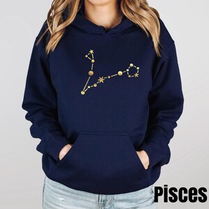 May include: A navy blue hoodie with a gold Pisces constellation design. The hoodie has a drawstring hood and a kangaroo pocket.