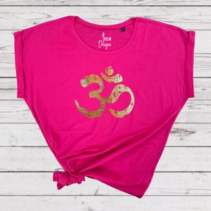 以下が含まれることがあります： ピンクのTシャツに、金色のオームのシンボルが前面にプリントされています。Tシャツはゆったりとしたフィット感で、袖はロールアップされています。Tシャツには「Inca Designs」の文字がプリントされています。