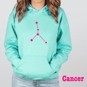 May include: A turquoise blue hoodie with a pink Cancer zodiac constellation graphic on the front. The word "Cancer" is printed in pink below the graphic.