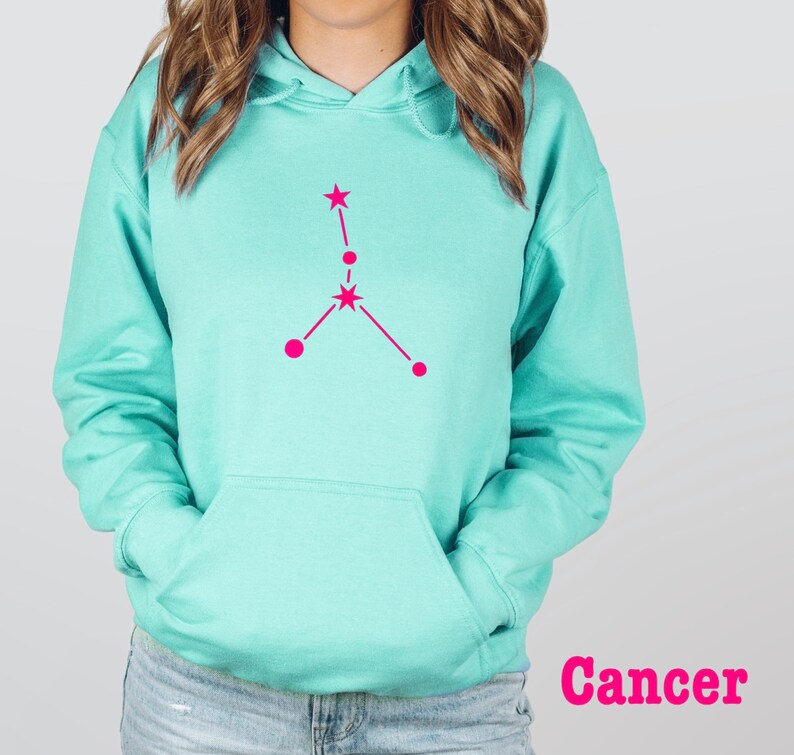 May include: A turquoise blue hoodie with a pink Cancer zodiac constellation graphic on the front. The word "Cancer" is printed in pink below the graphic.