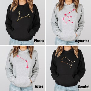 May include: Four gray and black hoodies with different zodiac constellation designs. The constellations are printed in gold and pink. The hoodies are all the same style, with a drawstring hood and a kangaroo pocket. The zodiac signs are Pisces, Aquarius, Aries, and Gemini.