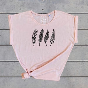 Może przedstawiać: Jasnor&oacute;żowy t-shirt z grafiką czterech czarnych pi&oacute;r z misternymi wzorami. Na przodzie koszulki nadrukowany jest tekst "Inca Designs".