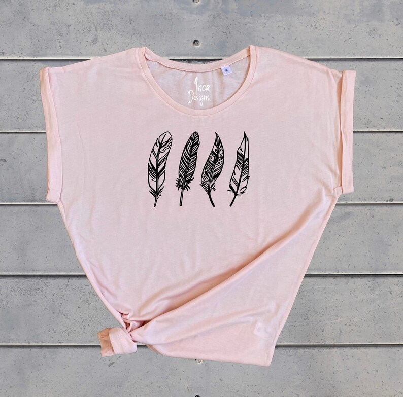 Może przedstawiać: Jasnor&oacute;żowy t-shirt z grafiką czterech czarnych pi&oacute;r z misternymi wzorami. Na przodzie koszulki nadrukowany jest tekst "Inca Designs".