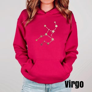 May include: A pink hoodie with a gold Virgo constellation design. The word "virgo" is printed in black at the bottom of the image.