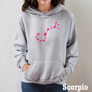 May include: Gray hooded sweatshirt with a pink outline of the Scorpio constellation. The text "Scorpio" is printed in black at the bottom of the image.