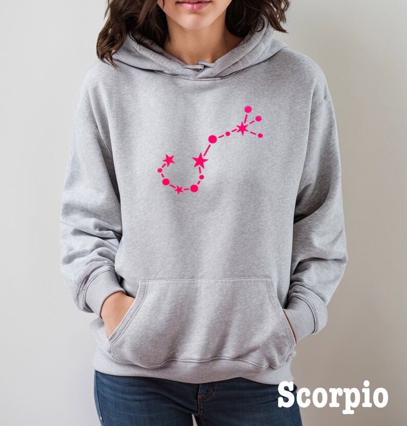 May include: Gray hooded sweatshirt with a pink outline of the Scorpio constellation. The text "Scorpio" is printed in black at the bottom of the image.