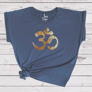 以下が含まれることがあります： 紺色のTシャツに、金色でオームのシンボルがプリントされています。オームのシンボルは、使い古したようなヴィンテージな外観です。Tシャツはゆったりとしたフィット感で、袖はロールアップされています。