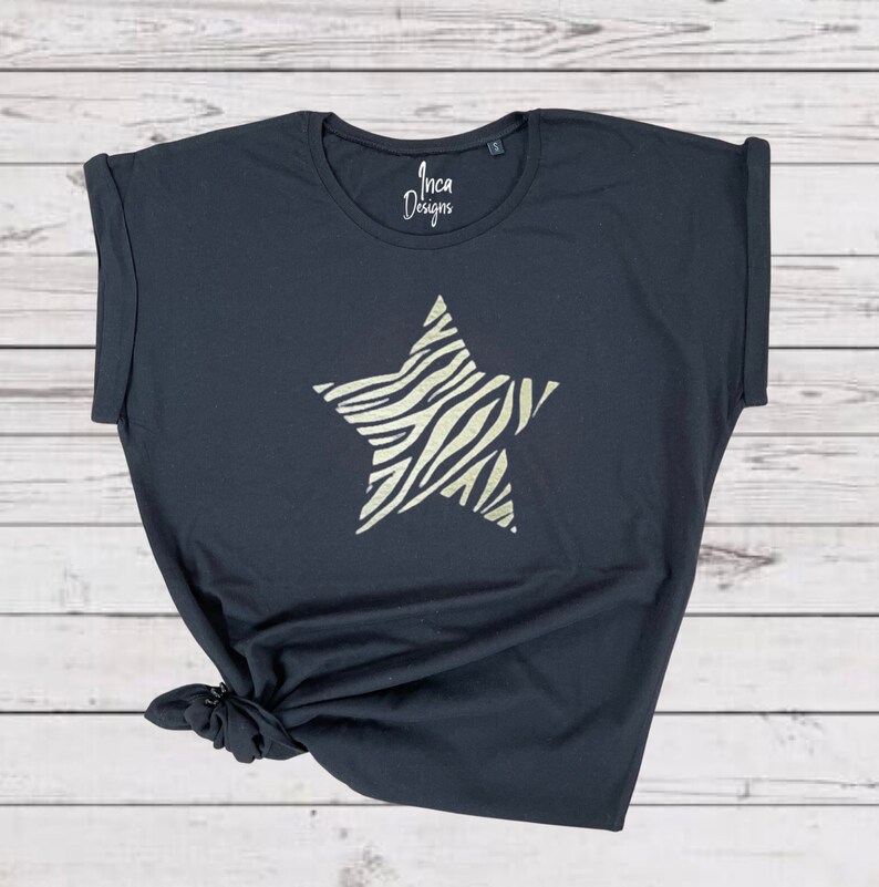 Pu&ograve; includere: Maglietta nera con una grande stella dorata stampata sul davanti. La stella ha un motivo a zebrato. La maglietta ha una manica arrotolata e dice "Inca Designs" sul davanti.