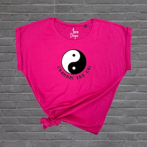 Könnte beinhalten: Ein pinkfarbenes T-Shirt mit einem schwarzen Yin-Yang-Symbol und dem Text "Channel the chi" auf der Vorderseite.