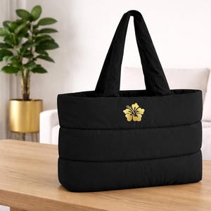 May include: Black quilted tote bag with a gold hibiscus flower emblem. The bag has two black handles and is sitting on a wooden table. The bag is a medium size and appears to be made of a soft, padded material.