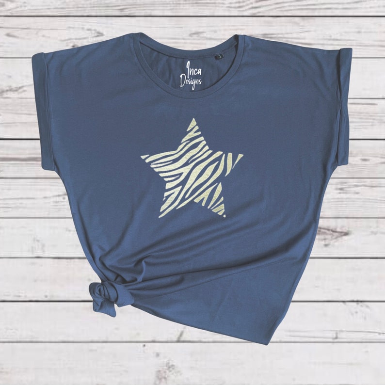 Pu&ograve; includere: Una t-shirt blu navy con un design a stella con stampa zebrata bianca e dorata. La t-shirt ha una vestibilit&agrave; rilassata e maniche arrotolate. Il testo "Inca Designs" &egrave; stampato sulla t-shirt.