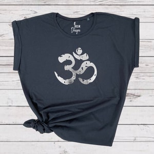 以下が含まれることがあります： 前面に銀色のメタリックなオームのシンボルがプリントされた黒いTシャツ。シャツはロールアップスリーブでゆったりとしたフィット感。シャツには「インカデザイン」の文字がプリントされています。