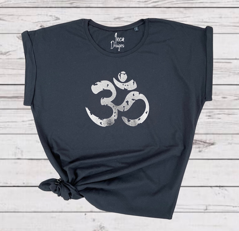 以下が含まれることがあります： 前面に銀色のメタリックなオームのシンボルがプリントされた黒いTシャツ。シャツはロールアップスリーブでゆったりとしたフィット感。シャツには「インカデザイン」の文字がプリントされています。