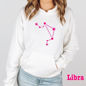May include: White hoodie with a pink Libra constellation graphic. The word "Libra" is printed in pink at the bottom of the image.