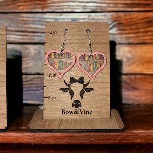 May include: A pair of pink heart-shaped earrings with a bookshelf design featuring colourful books. The earrings are displayed on a wooden stand with the text "Bow & Vine".