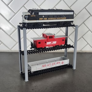 May include: Three toy train cars on a three-tiered metal stand. The top car is a black and white Norfolk Southern locomotive. The middle car is a red boxcar with the Norfolk Southern logo. The bottom car is a gray boxcar with the words "SOUTHERN" and "SR" on it.