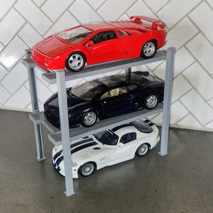 Puede incluir: Una estantería de metal de tres niveles con tres coches de juguete en ella. El coche de arriba es rojo, el del medio es negro y el de abajo es blanco.