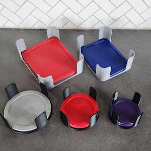 Może przedstawiać: Kolekcja pojemników i pokrywek do przechowywania Pyrex w różnych kolorach, w tym czerwonym, niebieskim, fioletowym i srebrnym. Pokrywki są prostokątne i okrągłe, a pojemniki są umieszczone w szarych i czarnych uchwytach.