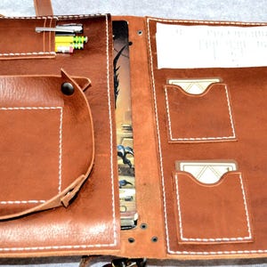 May include: Open brown leather gaming accessory with multiple pockets, a small tray, and pen holders. The accessory is stitched with white thread and has a strap for carrying. It contains cards, pencils, and paper.