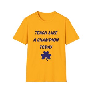 May include: Mustard yellow t-shirt with the text "TEACH LIKE A CHAMPION TODAY" in dark blue, along with a dark blue four-leaf clover graphic. The shirt has a crew neck and short sleeves.