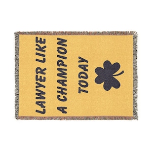 May include: Mustard yellow throw blanket with the text "LAWYER LIKE A CHAMPION TODAY" in navy blue, along with a navy blue shamrock. The blanket has a fringed edge.