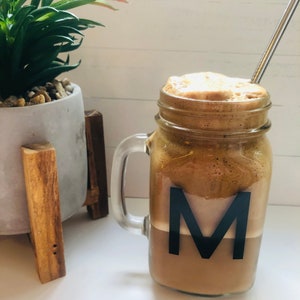 May include: A mason jar mug with a handle filled with a brown, frothy coffee drink. The drink has a layer of foam on top and a black letter "M" on the side of the jar.