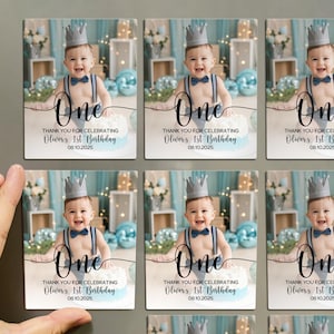 May include: Rectangular magnets featuring a baby wearing a crown and suspenders, with the word "One" in elegant script. The text reads "Thank you for celebrating Oliver's 1st Birthday 08.10.2025". The background includes blue balloons.