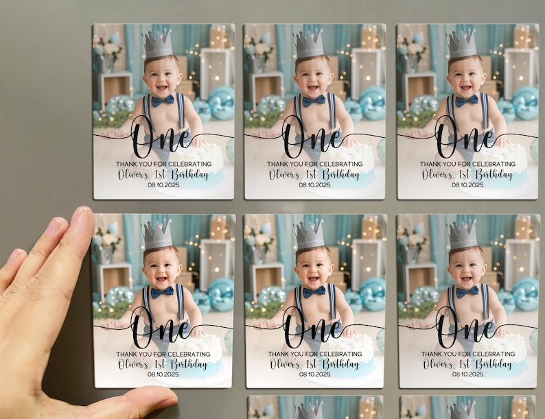 May include: Rectangular magnets featuring a baby wearing a crown and suspenders, with the word "One" in elegant script. The text reads "Thank you for celebrating Oliver's 1st Birthday 08.10.2025". The background includes blue balloons.