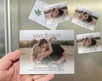 May include: Rectangular save-the-date magnets with a photo of a couple embracing. The magnets have the text "SAVE the DATE" and the names "Emma & James" along with the date and location. A green leaf-shaped card is also visible.
