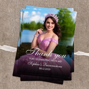May include: A stack of printed cards featuring a woman in a pink dress and crown. The cards have the text "Thank you for celebrating with me! Sophia's Quincea&ntilde;era 08.12.2025" in a script font. The background shows a lake and trees.