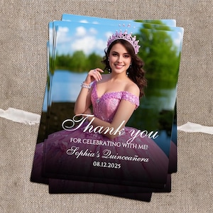 May include: A stack of printed cards featuring a woman in a pink dress and crown. The cards have the text "Thank you for celebrating with me! Sophia's Quincea&ntilde;era 08.12.2025" in a script font. The background shows a lake and trees.