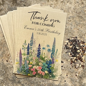 Puede incluir: Una pila de sobres de semillas de papel kraft con ilustraciones florales y el texto "Thank you for coming Emma's 50th Birthday 11.8.2025". Un peque&ntilde;o mont&oacute;n de semillas est&aacute; al lado de los sobres.
