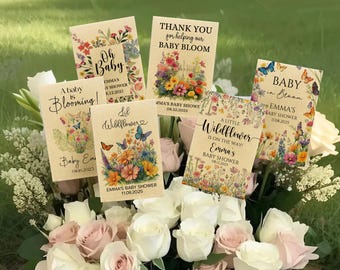 May include: A collection of baby shower seed packets with floral and butterfly designs. Each packet features text such as "Oh Baby," "A baby is Blooming!" and "Thank you for helping our baby bloom." The packets are arranged in a bouquet of white and pink roses.