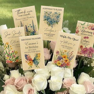 Puede incluir: Una colecci&oacute;n de recuerdos de paquetes de semillas personalizados con dise&ntilde;os florales y texto. Los paquetes presentan frases como "Gracias", "Birthday Blooms" y nombres con fechas de cumplea&ntilde;os. Los paquetes est&aacute;n dispuestos en un ramo de rosas blancas y rosas.