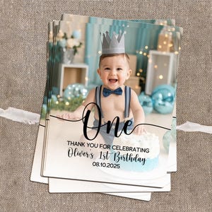 May include: A stack of photo cards featuring a baby wearing a crown and bow tie, with the word "One" and birthday details. The cards have a light blue and white color scheme, with a cake in the image.