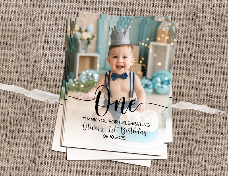 May include: A stack of photo cards featuring a baby wearing a crown and bow tie, with the word "One" and birthday details. The cards have a light blue and white color scheme, with a cake in the image.