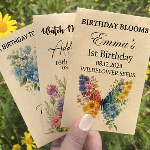 Puede incluir: Tres sobres de semillas con dise&ntilde;os florales y texto. Uno dice "A Birthday To", otro "Watch Me Add" y el tercero "Birthday Blooms Emma's 1st Birthday 08.12.2025 Wildflower Seeds". Los sobres presentan ilustraciones de mariposas azules, rosas y coloridas.